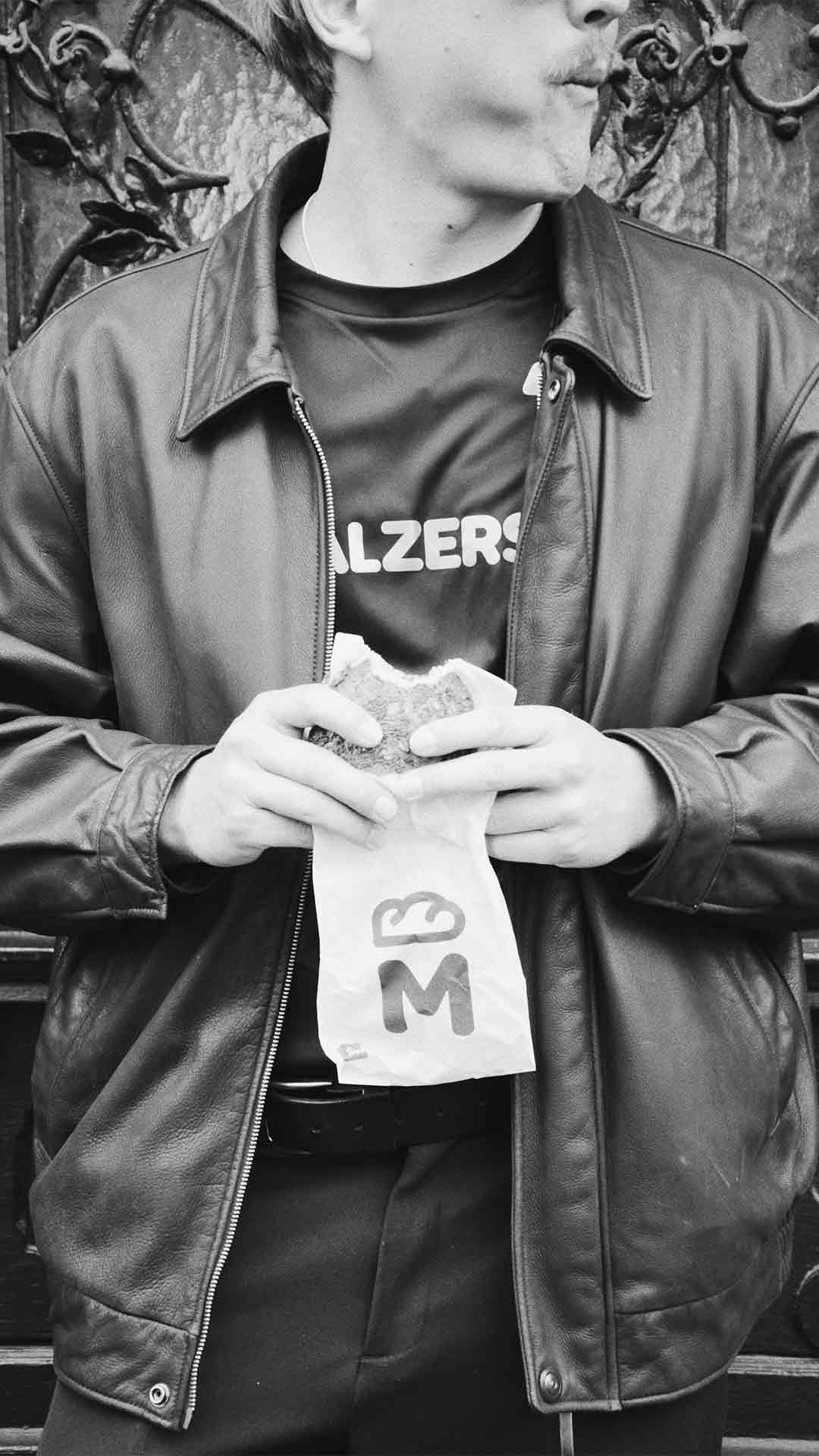 Person mit Malzers Shirt und Ledermantel hält ein belegtes Brot in der Hand
