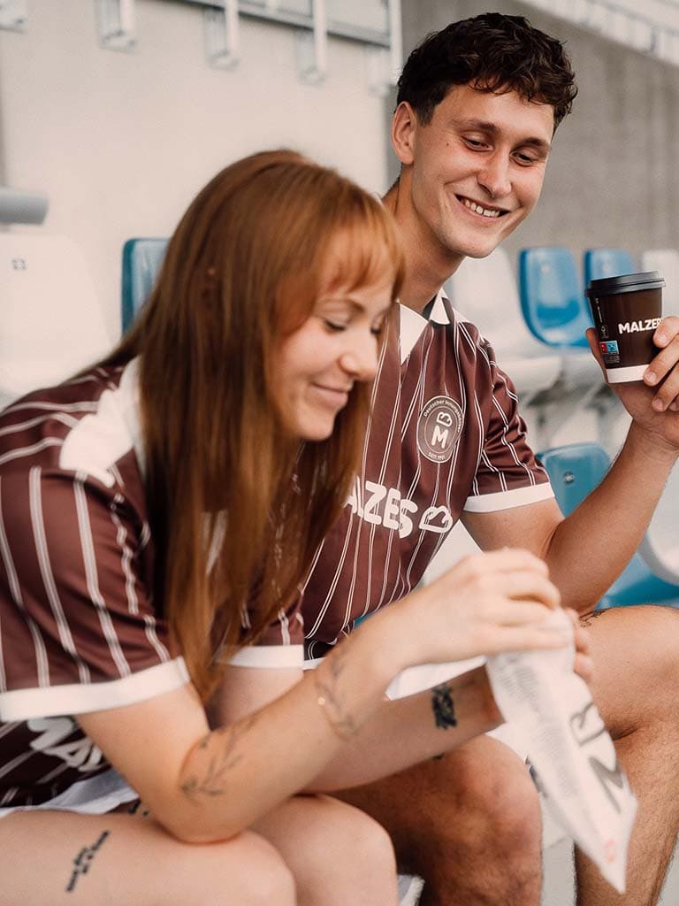 Zwei Personen im Malzers Running Jersey auf einer Stadiontribüne mit Malzers Kaffeebecher